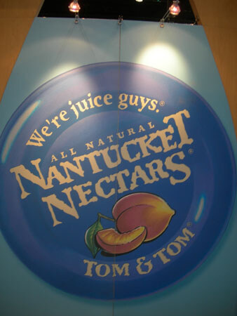 A large round Nantucket Nectars sign reading We're juice guys, All Natural, Tom & Tom, with a peach illustration on a blue background.
