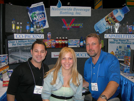 Two men and a blonde woman pose at the Nationwide Beverage Bottling, Inc. booth, with signs for co-packing, Vuzion Enhanced Water and Wet Mist and bottled water displays behind them.