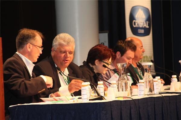 Five people sit at a draped panel table with microphones, water carafes and small bottles. One man in glasses speaks, and a Crystal banner hangs in the background.