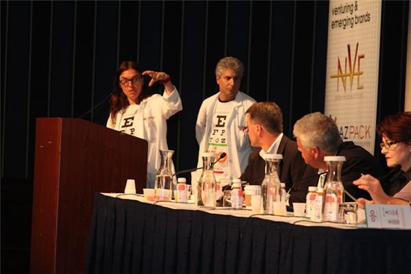 Two people in white lab coats with eye-chart lettering stand near a podium, and one gestures. Panelists sit at a table with bottles. An NVE banner is at right.