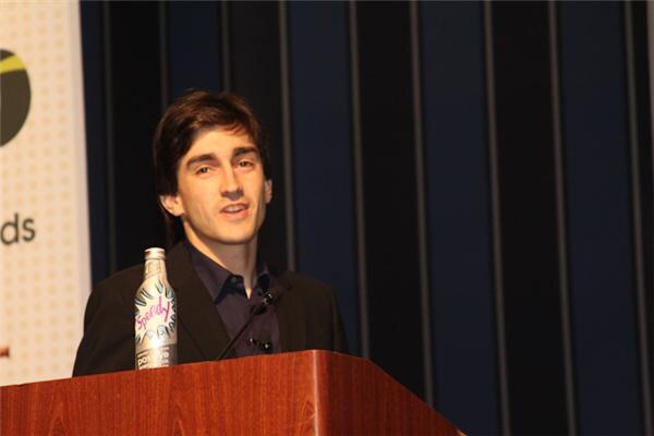 A young man with dark hair speaks at a wooden podium with a microphone. A Speedy bottle stands on the podium, and a dark curtain is behind him.