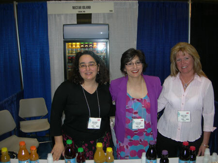 Three smiling women with name badges stand behind a table of juice bottles at the Nectar Island booth, with a lit refrigerator display behind them.