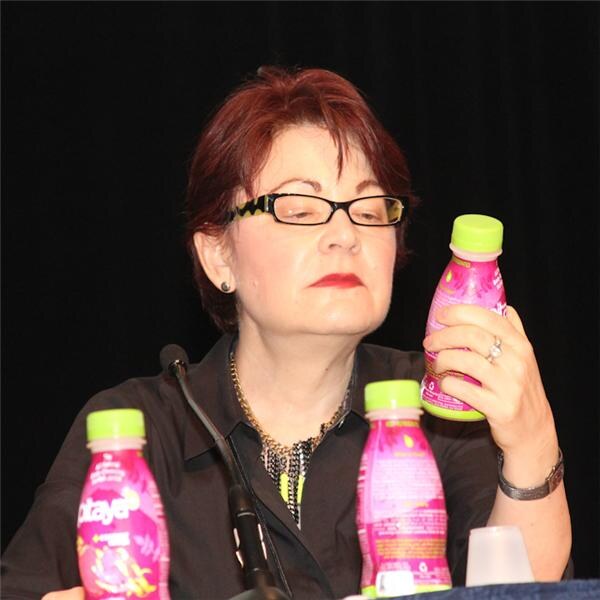 A woman with short red hair and black-framed glasses looks at a pink bottle with a green cap she holds up, with two more pink Datayer bottles on the table in front of her.
