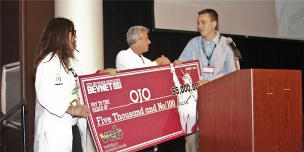 A woman in a white lab coat, a man in a white shirt and a man at a podium with a microphone hold a large check made out to OJO for $5,000 from veb and BevNET Live.