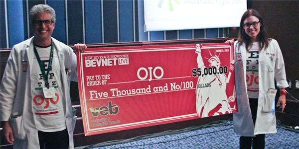 A man and a woman in white lab coats with OJO shirts stand on either side of a large red check for $5,000 made out to OJO, with BevNET Live and veb logos.