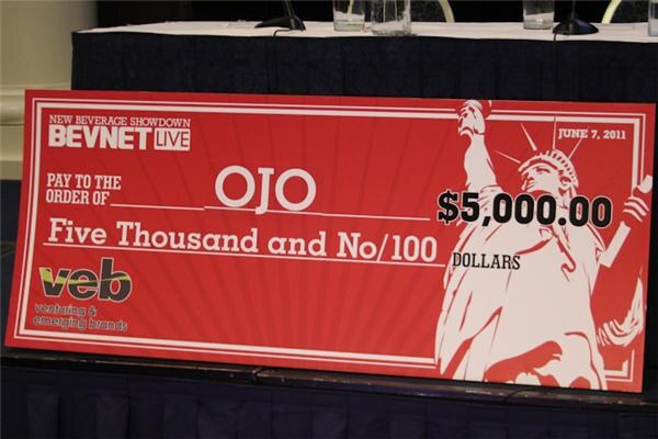 A large red oversized check dated June 7, 2011, made out to OJO for $5,000.00, with BevNET Live New Beverage Showdown and veb logos and a Statue of Liberty graphic.