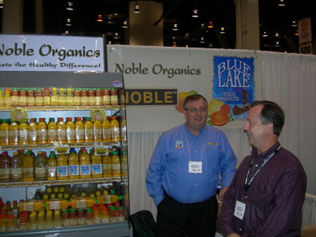 Two men with name badges stand at the Noble Organics booth beside a refrigerated case of juice bottles, with a Blue Lake Citrus Products sign behind them.