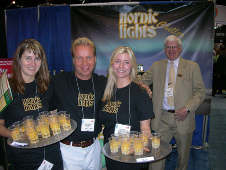 Two women and a man in black Nordic Lights shirts hold trays of small drinks, with an older man in a tan suit beside them, in front of a Nordic Lights banner.