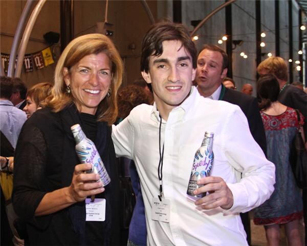 A smiling woman and a young man in a white shirt, both wearing name badges, each hold a Speedy bottle while standing in a crowd at an evening event with string lights.