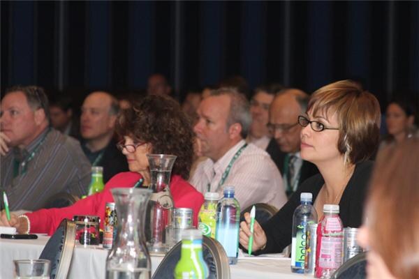 Seated attendees with lanyards watch a presentation, holding pens, with water carafes and bottles including Balance Water on the tables in front of them.