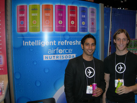 Two smiling men in black stand in front of a blue Nutrisoda Airforce display showing seven colorful cans and the words Intelligent refreshment. One holds a pink can, the other a green can.