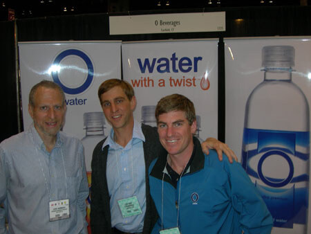 Three men stand close together in front of O Water banners reading water with a twist, beneath an O Beverages sign. Two wear conference badges.