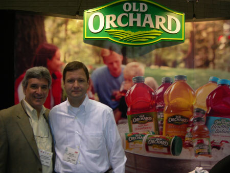 Two men stand in front of an Old Orchard sign and a display of Old Orchard juice bottles and cans in red and orange. One wears a tan jacket, the other a white shirt.