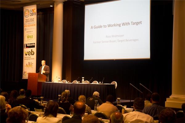 A speaker stands at a podium in a ballroom beside a large screen reading A Guide to Working With Target, while seated attendees face the stage.