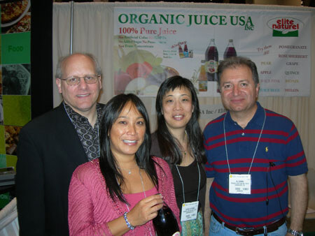 Four people pose together in front of an Organic Juice USA and Elite Naturel banner listing juice flavors; one woman holds a dark bottle.