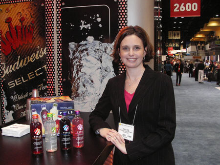 A smiling woman in a dark pinstriped jacket with a name badge stands beside a counter holding Peels bottles and a variety pack, with Budweiser Select signage behind her and the show floor in the background.