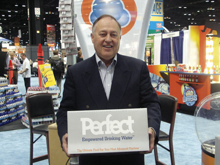 A smiling man in a dark jacket holds a white box labeled Perfect Empowered Drinking Water in an aisle of a trade show hall with other booths behind him.