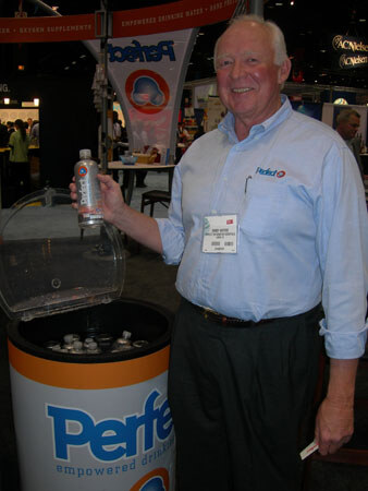 A smiling white-haired man in a light blue shirt holds a bottle of water beside an open Perfect cooler display. A Perfect banner hangs overhead and his shirt carries a Relief logo.