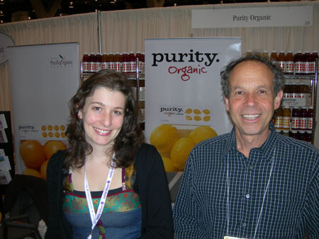 A smiling woman with dark curly hair and a smiling man in a checkered shirt stand in front of Purity Organic banners with oranges and shelves of bottled juices.