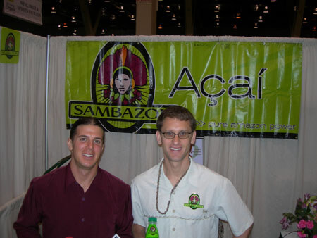Two smiling men stand in front of a green Sambazon Açaí banner. One wears a maroon shirt, the other a light shirt, glasses and a necklace, with a green bottle in front of him.