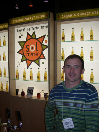 A man in a blue and green striped sweater and a name badge stands in front of lit shelves of bottles under Sol Mate signs reading 'Sparkling Yerba Mate' and 'Organic Energy Drink'.