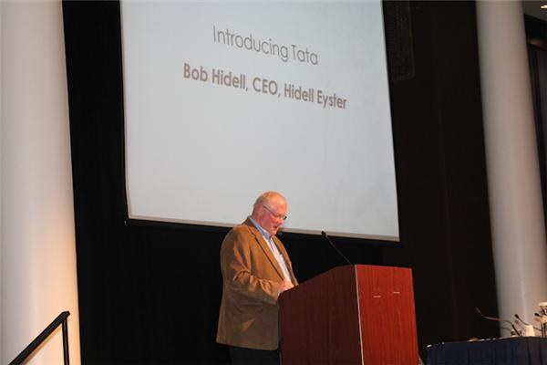 Bob Hidell of Hidell Eyster, in a tan jacket and glasses, stands at a wooden podium. A projected slide behind him reads Introducing Tata, Bob Hidell, CEO, Hidell Eyster.
