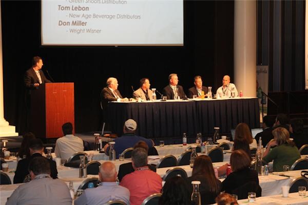 Five panelists sit at a draped table facing an audience while a man stands at a podium at left. A slide lists Green Shoots Distribution, New Age Beverage Distributors and Wright Wisner.