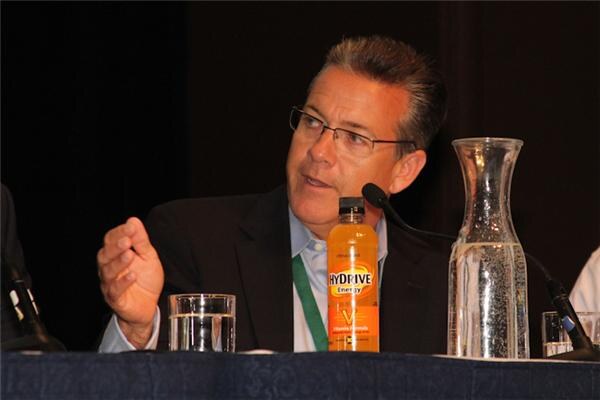 A man with glasses and a green lanyard sits at a panel table and gestures with one hand while speaking. A bottle of HyDrive Energy, a water glass and a carafe sit on the table in front of him.