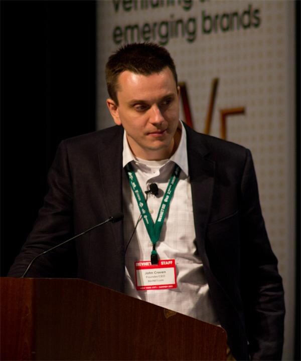 John Craven of BevNET.com, wearing a dark jacket and a green staff lanyard with a name badge, stands at a wooden podium with microphones in front of a banner.