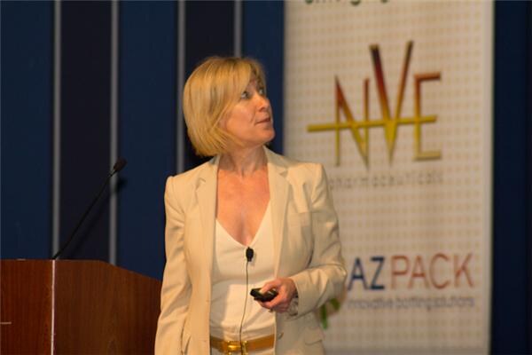 A blonde woman in a cream blazer and white top stands at a podium holding a small remote and looks up and to the side. Behind her is a banner for NVE Pharmaceuticals and AZ Pack.