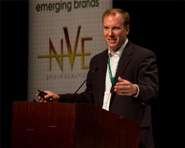 A smiling man in a dark jacket and green lanyard stands at a podium holding a small clicker and gesturing. Behind him is a banner reading emerging brands and NVE Pharmaceuticals.