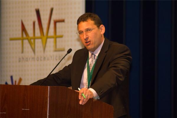 A man with glasses, a dark suit and a green lanyard speaks at a wooden podium with a microphone. A banner for NVE Pharmaceuticals is behind him.