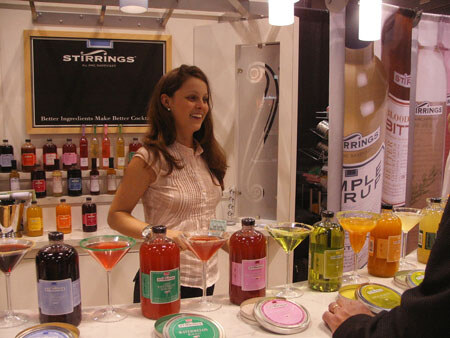 A smiling woman stands behind a counter at the Stirrings booth, with cocktail glasses, bottles of colored mixers and a Stirrings sign behind her.