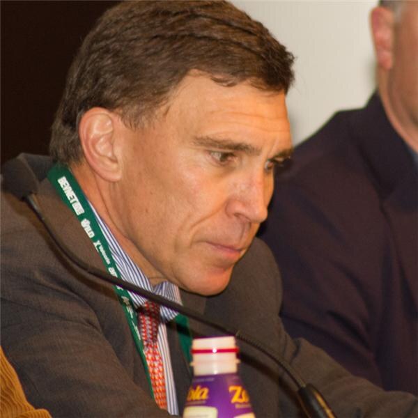 Close-up of a man with a green lanyard and striped tie looking downward while seated at a table with a microphone. A Zola bottle sits in the foreground and another person is partly visible behind him.