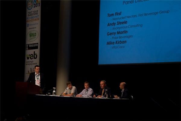A man stands at a podium at left while four panelists sit at a table. A screen lists Nantucket Nectars, First Beverage Group, Anonymous Consulting, Polar Beverages and Vita Coco.