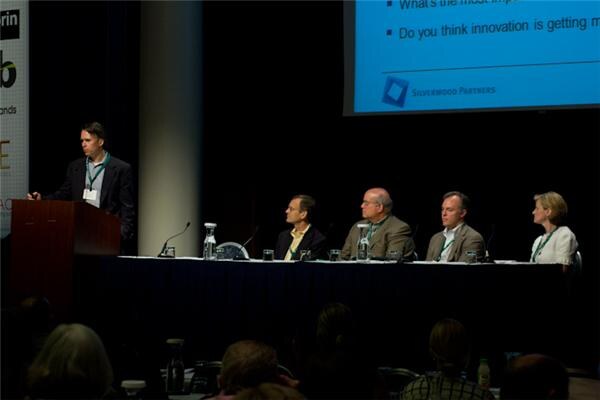 A man stands at a podium beside a long panel table where four people sit. A projected slide with the Silverwood Partners logo shows discussion questions above them.