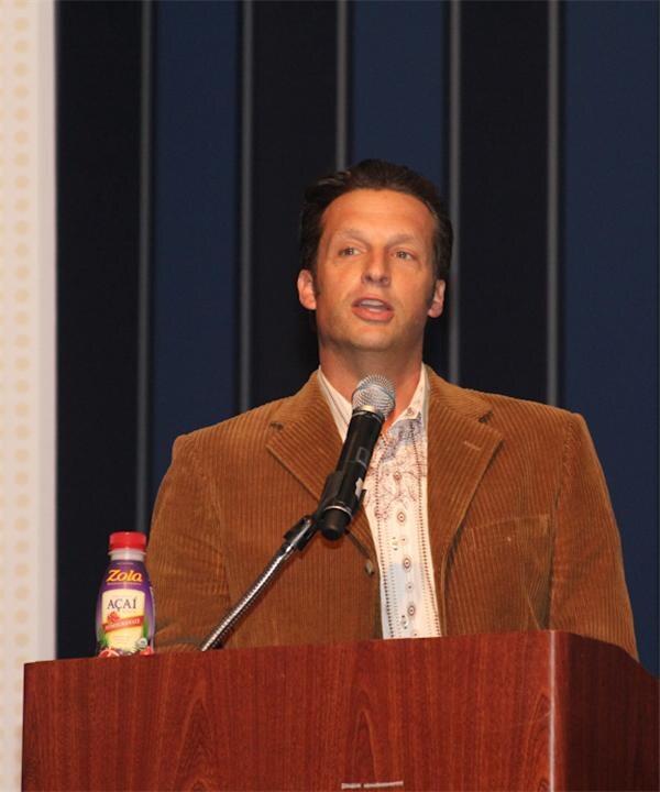 A man in a brown corduroy jacket speaks into a microphone at a wooden podium. A bottle of Zola Acai juice sits at the lower left of the podium.