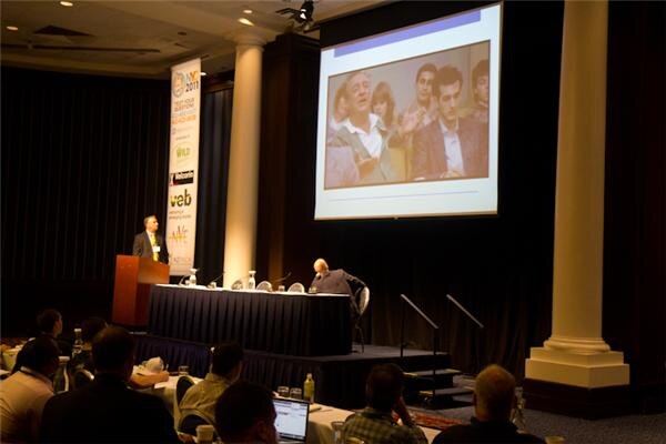 A man stands at a wooden podium beside a tall veb banner on a stage. A large projection screen shows a photo of several people. A seated person is at a draped table; attendees sit in the foreground.