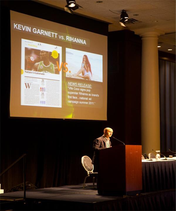 A man speaks at a podium on stage beneath a large projected slide titled Kevin Garnett vs. Rihanna, with a news release about Vita Coco.