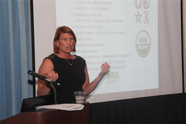 A woman with short reddish hair in a black sleeveless top speaks at a podium and gestures with both hands. A slide behind her shows certification logos, USDA Organic and the Wild logo.