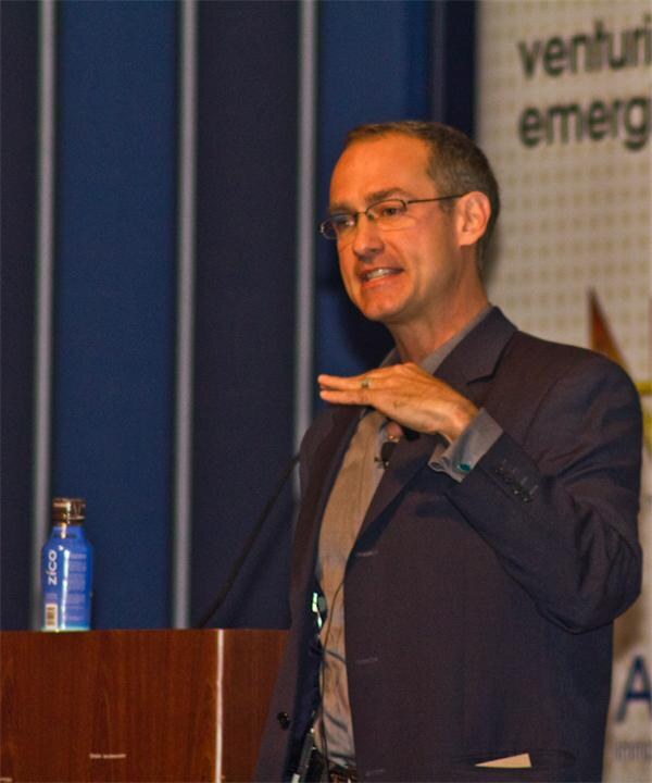 A man with glasses in a dark blazer gestures while speaking at a wooden podium, with a blue Zico bottle on the podium and a banner behind him.