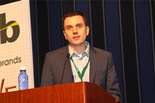 A man in a gray blazer and green lanyard speaks into a microphone at a wooden podium, with a ZICO bottle beside him and a veb banner behind.