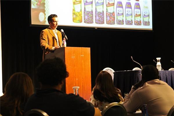 A man in a brown jacket stands at a podium on stage while a screen above shows a row of Zola Acai bottles. Audience members are seen from behind in the foreground.