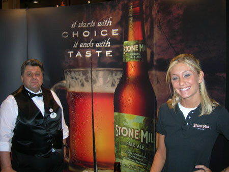 A man in a black vest and bow tie and a smiling blonde woman in a dark polo stand beside a large Stone Mill Pale Ale bottle graphic reading 'it starts with choice, it ends with taste'.