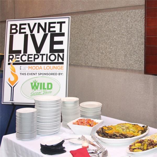 A sign reads BevNET Live Reception, Moda Lounge, sponsored by Wild. Stacks of plates and trays of food sit on a draped table beside it.