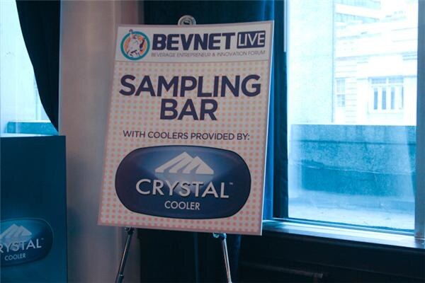 An easel sign reads BevNET Live Sampling Bar with coolers provided by Crystal Cooler. A Crystal Cooler unit stands at the left edge next to a window.