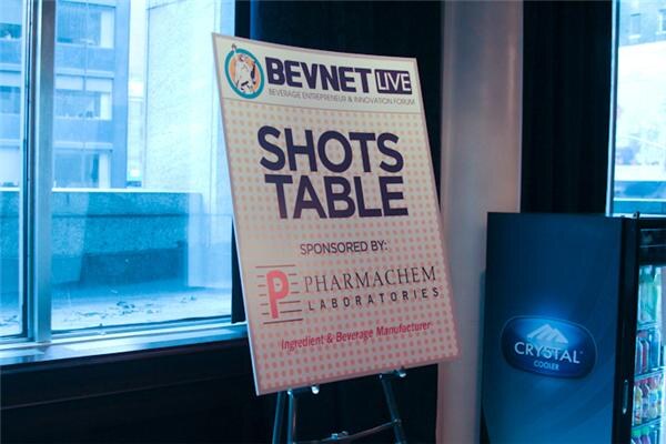 An easel sign reads BevNET Live Shots Table, sponsored by Pharmachem Laboratories. A Crystal Cooler beverage cooler stands to the right beside a window.