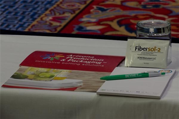 On a white-draped table sit an Arizona Production & Packaging brochure, a Fibersol-2 ADM/Matsutani LLC packet leaning on a glass, and a BevNET.com pen on a notepad.