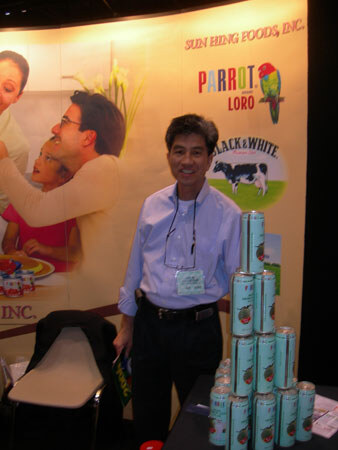 A man in a light blue shirt stands beside a stack of canned drinks at the Sun Hing Foods booth, in front of Parrot and Black & White logos and a family photo backdrop.