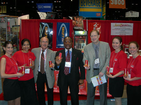 Three men in business attire and four women in red shirts stand in a row holding small red cans in a booth with Tantra, Nestlé and Bounty signs and red drapes behind them.